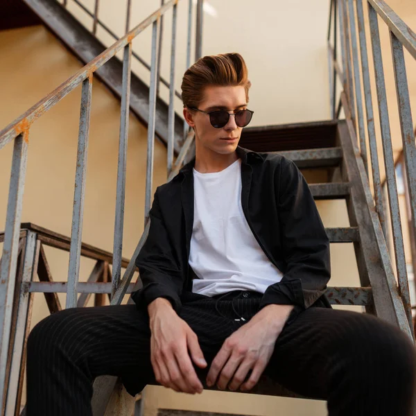 Guapo joven hipster hombre con peinado en ropa de moda en gafas de sol con estilo posa sentado en una escalera de hierro vintage al aire libre. Un hombre europeo moderno descansando en la ciudad. Moda callejera . — Foto de Stock