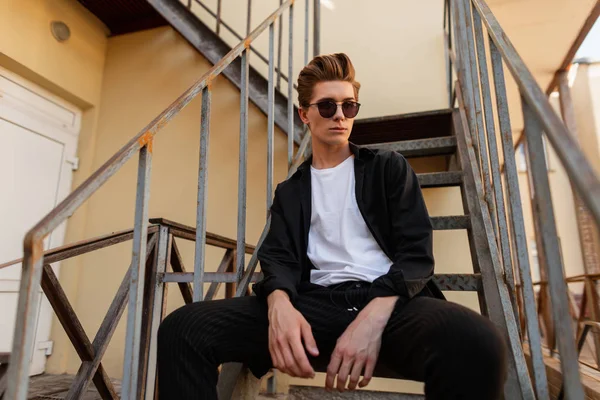 Demir merdivenlerde oturan zarif genç hipster adam. Bir yaz günü açık havada şık bir saç stili ile trendy güneş şık giysiler çekici adam modeli. Şık erkek giyim.