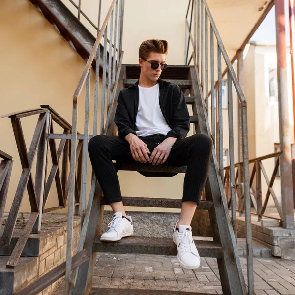 Elegante hombre hipster joven en gafas de sol de moda en una camisa negra en una camiseta con elegantes pantalones a rayas en zapatillas de deporte se sienta en una escalera de hierro vintage en la calle. Hombre europeo está descansando en la ciudad . — Foto de Stock