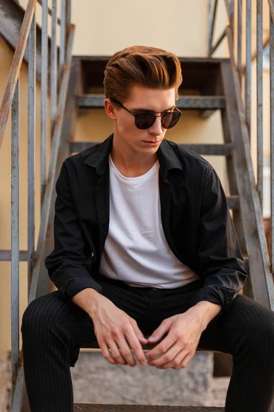 Mladý pohledný hipíček v módních slunečních brýlích s elegantním účesu ve starožitných černých košili v bílých tričku v elegančních kalhotách sedí na železném schodišti na ulici. Model moderního módního muže. — Stock fotografie