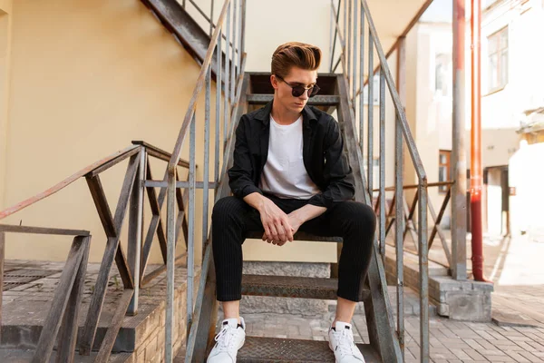 Moderne jonge hipster man in trendy zonnebril in stijlvolle zwarte shirt in een t-shirt in elegante gestreepte broek in Sneakers zit op een ijzeren oude trap in de straat. Coole kerel rust in de stad. — Stockfoto