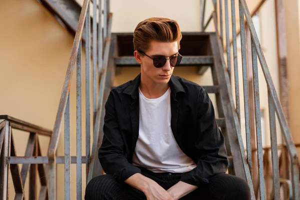 El joven estadounidense hipster con una camisa elegante en una camiseta con gafas de sol de moda se sienta en una escalera de hierro en la ciudad. Chico guapo con un peinado elegante descansando en la calle. Moda masculina —  Fotos de Stock