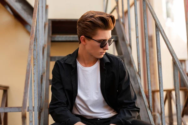 Joven hipster guapo en gafas de sol de moda con un peinado de moda en una elegante camisa negra en una camiseta blanca sentada en una escalera de hierro en la ciudad. Un tipo atractivo. Ropa de hombre con estilo . — Foto de Stock
