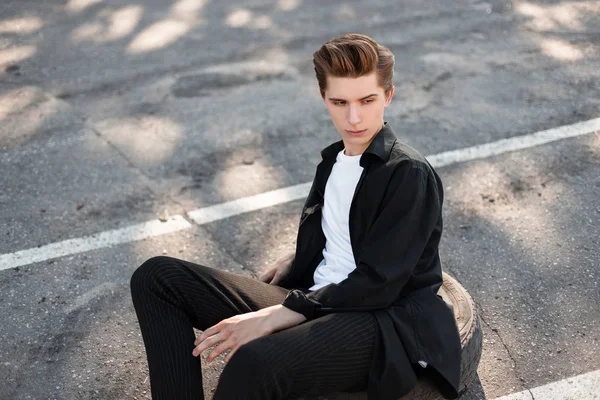 Atractivo joven con un peinado elegante en una camisa elegante en pantalones de rayas vintage en zapatillas blancas de moda se sienta en el asfalto en la calle. Un tipo urbano descansa en la ciudad en un día soleado . — Foto de Stock