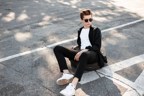 Ein junger Hipster in schwarzer modischer Kleidung mit trendiger Sonnenbrille und stylischen weißen Turnschuhen sitzt auf dem Asphalt in der Stadt. schöner Kerl, der sich an einem sonnigen Sommertag im Freien ausruht. Straßenstil. — Stockfoto