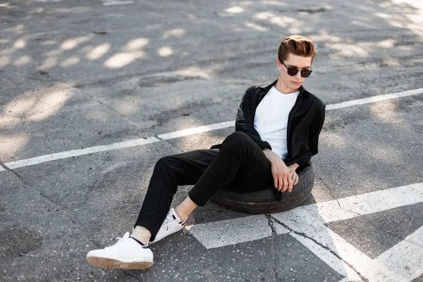 Joven y elegante hombre hipster con peinado de moda en una elegante camisa negra con pantalones a rayas en zapatillas blancas sentadas en una rueda de goma en el pavimento. chico urbano en gafas de sol está descansando en el sol . — Foto de Stock