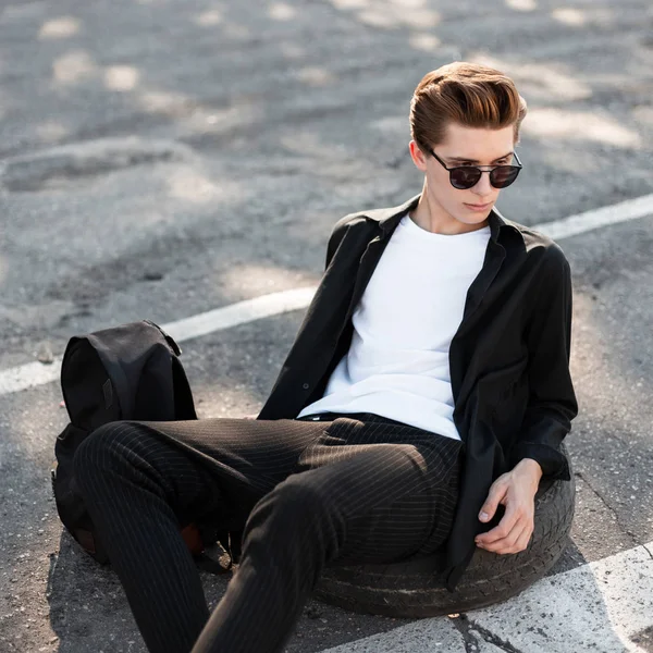 Elegante joven hipster hombre en gafas de sol en ropa de moda con un peinado elegante se relaja sentado en la rueda de goma del coche en una calle en el día soleado. Hombre urbano descansando en la calle . — Foto de Stock