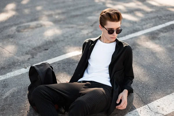 Knappe jonge man met trendy kapsel ontspant op asfalt op een zonnige dag. Modieuze hipster kerel in stijlvolle kleding met een rugzak is rusten op de straat en genieten van de zon. Street fashion. — Stockfoto