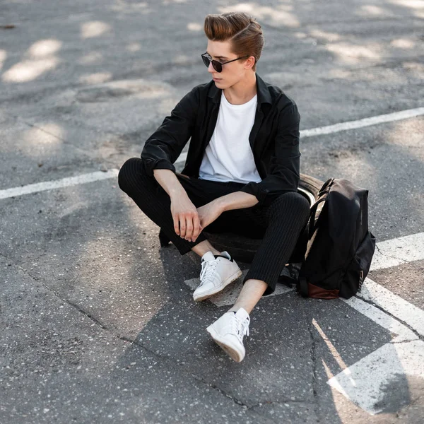 Homem moderno jovem hipster em óculos escuros em roupas da moda em sapatos brancos relaxa sentado na roda de borracha do carro em uma rua no dia ensolarado. Modelo cara legal descansando na rua . — Fotografia de Stock