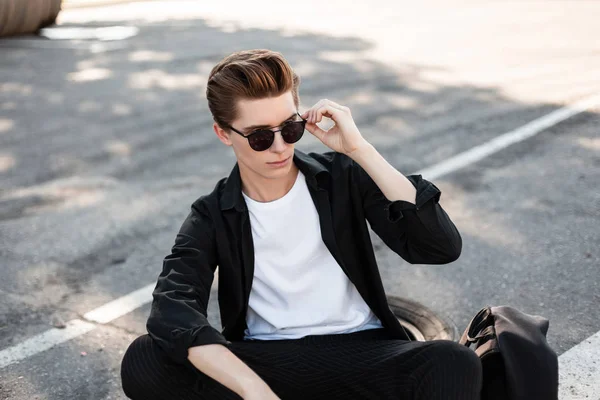 Joven hombre hipster con estilo en gafas de sol de moda en una camisa elegante en pantalones a rayas descansando en un día soleado al aire libre. Guapo chico modelo disfruta soleado verano día sentado en el asfalto en la ciudad . — Foto de Stock