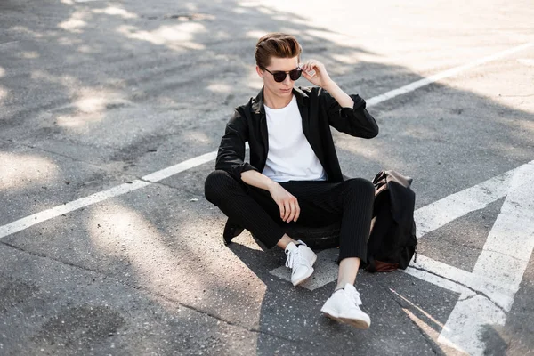 Homem hipster jovem urbano com penteado em óculos de sol em uma camisa preta em calças listradas na moda em tênis com uma mochila sentar-se em uma roda de borracha em uma rua em um dia ensolarado. Um tipo bonito. Estilo de rua . — Fotografia de Stock