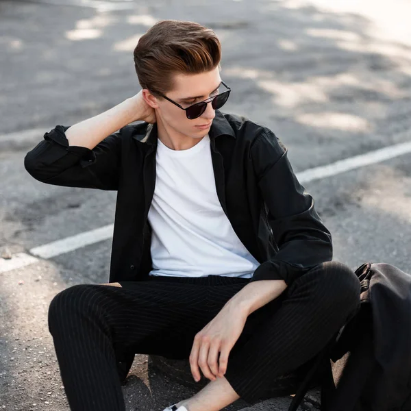Joven hombre hipster guapo con peinado elegante en gafas de sol en una camisa en pantalones a rayas con una mochila negra sentarse en el asfalto en la ciudad. chico atractivo es relajarse en la calle en un día soleado —  Fotos de Stock