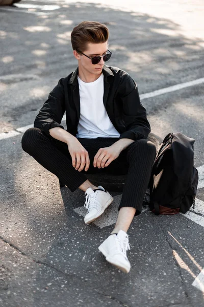 Jovem atraente em uma camisa elegante em óculos de sol elegantes em calças com uma mochila em tênis da moda senta-se no asfalto na rua. Bonito hipster cara relaxa na cidade em um dia ensolarado . — Fotografia de Stock