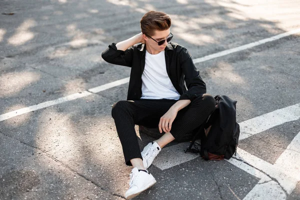 Nice young man hipster in elegant black clothes in white trendy sneakers with a backpack in sunglasses sits on a rubber wheel on the street. Cool guy relaxes in the sun outdoors on a summer day.