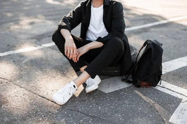Κοντινό-up των ανδρών μοντέρνα πόδια. Νέος άνθρωπος της μόδας σε κομψό μαύρο πουκάμισο σε ριγέ vintage παντελόνια σε λευκά αθλητικά παπούτσια με κομψό μαύρο σακίδιο κάθεται στην άσφαλτο... συλλογή από μοντέρνα Ανδρικά. — Φωτογραφία Αρχείου