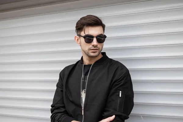 Nice attractive young hipster man with stylish hairstyle in fashionable black clothes in trendy dark sunglasses is resting near a white vintage metallic wall in the street. Cool guy. American style. — Stock Photo, Image