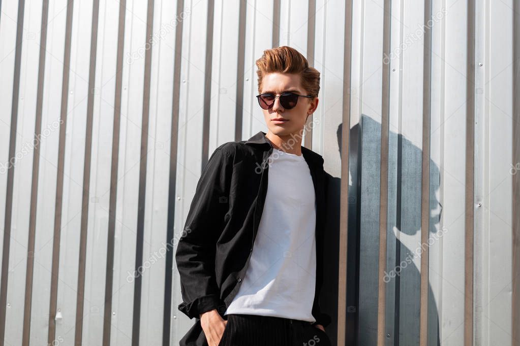 Young cool hipster man with a trendy hairstyle in elegant clothes in fashionable dark sunglasses poses near a silver metal wall in the street on a sunny summer day. Modern urban guy enjoys the sun. 