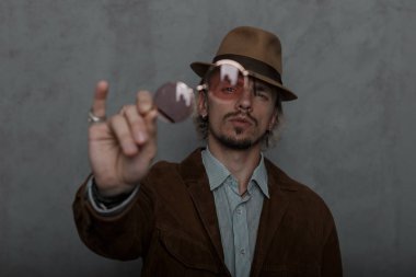 Genç acımasız hipster adam kamera trendy kırmızı yuvarlak gözlük gösteriyor. Zarif retro şapka vintage giysiler moda adam modeli odada gri bir duvarın arka planda pozlar. Close.