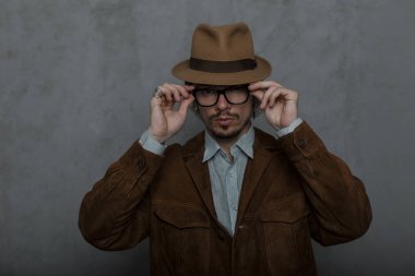 Klasik bir gömlek zarif kahverengi ceket bir retro şapka sakallı yakışıklı genç adam hipster stüdyoda gri bir duvarın yanında duran trendy gözlük kapalı alır. Çekici eski moda adam. 