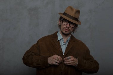 Klasik bir gömlek düğmeleri stüdyoda gri bir duvar yakınında şık kahverengi ceket şık şapka trendy yuvarlak gözlük Brutal genç sakallı hipster adam. Eski moda adam model. Retro tarzı