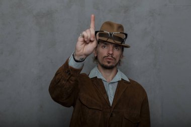 Yakışıklı adam hipster dikkat çeken onun işaret parmağını yükseltir. Odada gri bir duvarın yanında poz şık kahverengi ceket yuvarlak gözlük bir vintage şapka sakallı güçlü genç adam modeli.