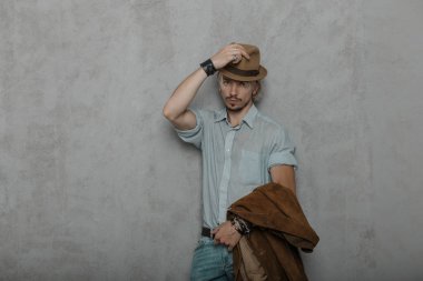 Deri bilezik standları ile kot pantolon bir klasik gömlek kahverengi ceket ile sakallı eski moda genç adam hipster duruyor ve gri duvarın yakınındaki odada şık şapka ayarlar. Yakışıklı adam modeli.