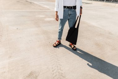 Siyah kumaş çanta ile şık bir gömlek moda mavi kot genç kentsel adam gri bir binanın yanında duruyor. Erkek giyim yeni moda yaz koleksiyonu. Close.