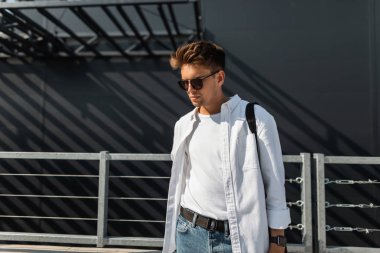 Siyah bir çanta koyu güneş gözlüğü içinde mavi kot şık beyaz giysiler içinde Yakışıklı kentsel genç hipster adam şehirde modern bir binanın yakınında duruyor. Güneşli bir günde modern trendy adam açık havada.