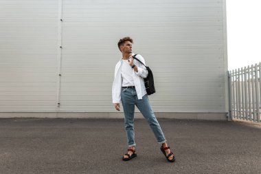 Siyah bir çanta ile deri kırmızı sandalet şık giysiler Içinde Avrupalı genç adam modeli şehirde beyaz modern binanın yakınında dinleniyor. Yakışıklı şehirli adam. Moda yazlık erkek giyim. Trendy görünüm.