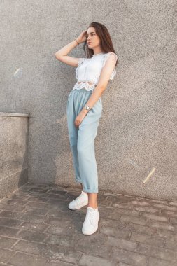 Deri beyaz spor ayakkabı şık bir lacy t-shirt zarif pantolon genç sevimli kadın şehirde bir vintage gri binanın yakınında duruyor. Yaz güneşli bir günde bir yürüyüş modern kız model.