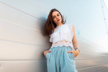 Şık mavi pantolon şık bir beyaz dantel bluz zarif modern genç model kadın şehirde bir vintage beyaz duvar yakınında pozlar. Avrupa seksi kız açık havada rahatlatır. Trendy yaz görünümü.