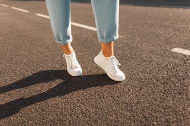 Şık mevsimlik kadın ayakkabıları. Parlak bir yaz güneşli bir günde asfalt üzerinde deri beyaz trendy spor ayakkabı mavi zarif pantolon kadın bacaklarının closeup. Modern kız yolda yürüyor.