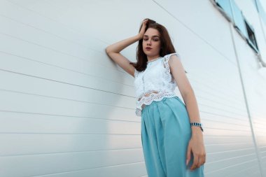 Şık mavi pantolon moda dantel bluz Avrupa kentsel genç esmer kadın beyaz bir vintage duvar yakınında açık havada dinlenir. Güzel kız bir yaz gününde açık havada rahatlar. Yeni koleksiyon.
