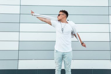 Yaz beyaz ve siyah moda güneş gözlüğü kot giysiler genç şık hipster adam modern bir binanın yakınında pozlar açık. Amerikalı yakışıklı adam şehirde dolaşıyor. Sokak stili. Erkek giyim.