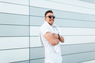 Şehirde modern bir binanın yakınında poz moda saç modeli ile şık güneş gözlüğü şık bir beyaz t-shirt Mutlu yakışıklı genç hipster adam. Dışarıda gülümseyen neşeli adam. Yaz stili.