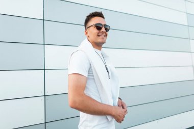 Bir yaz gününde şehirde modern bir çizgili binanın yakınında boyun bir madalyon ile şık bir beyaz t-shirt güneş gözlüğü neşeli genç hipster adam duruyor. Mutlu iyi adam modeli. Sokak modası