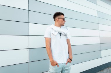 Şık bir beyaz t-shirt mavi vintage kot güneş gözlüğü Genç çekici adam hipster modern bir çizgili duvar yakınında şehirde dinleniyor. Bir yaz gününde açık havada saç modeli ile Yakışıklı adam.