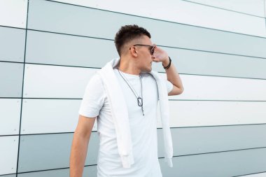 Şehirde modern bir binanın yakınında poz moda saç modeli ile şık güneş gözlüğü şık bir beyaz t-shirt Kentsel yakışıklı genç hipster adam. Çekici adam modeli açık havada. Yaz stili.