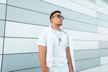 Bir yaz gününde şehirde modern bir çizgili binanın yakınında boyun bir madalyon ile şık bir beyaz t-shirt güneş gözlüğü şık genç hipster adam duruyor. Çekici erkek modeli. Sokak modası.