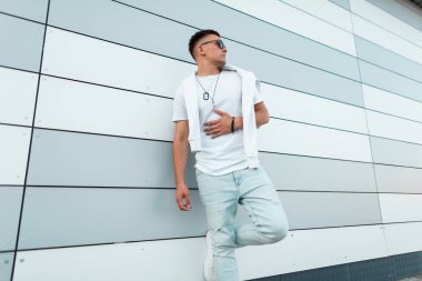 Siyah güneş gözlüğü siyah moda yaz beyaz ve kot giysiler genç şık adam hipster şehirde modern bir duvar yakınında açık havada rahatlatır. Sokakta dinlenen çekici manken. Erkek giyim.