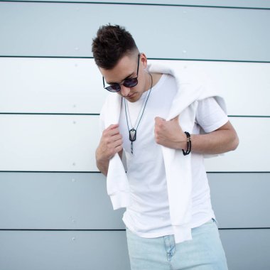 Şehirde modern bir binanın yakınında poz moda saç modeli ile şık güneş gözlüğü trendy beyaz t-shirt Yakışıklı güzel genç hipster adam. Kentsel adam model açık havada. Yaz stili.