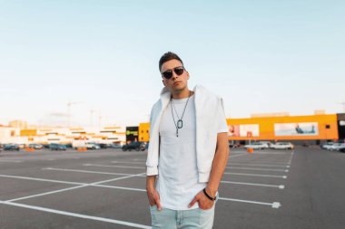 Şık bir saç modeli ile şık siyah güneş gözlüğü şık beyaz giysiler içinde Güzel genç adam hipster bir şehir park parlak güneşli bir günde duruyor. Urban American guy hipster. Yaz stili.
