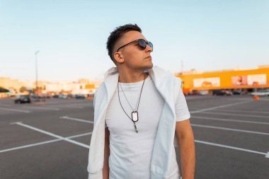 Şık bir saç modeli ile şık siyah güneş gözlüğü şık beyaz giysiler yakışıklı modern genç adam bir şehir otoparkparlak güneşli bir günde duruyor . Amerikalı hipster. Yaz stili.