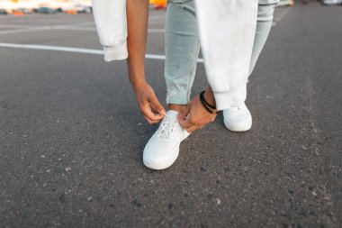 Şık genç adam kaldırımda duruyor ve bir yaz gününde deri beyaz spor ayakkabıları üzerinde ayakkabı bağcıkları bağlama. Erkek bacakları yakın çekim. Moda erkek ayakkabıları yeni mevsimlik koleksiyonu.