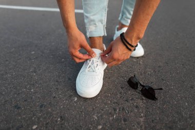 Deri beyaz trendy ayakkabı mavi kot genç şık adam güneş gözlüğü yanında yolda duruyor ve ayakkabı bağcıkları düzelten. Moda erkek spor ayakkabıları ve aksesuarları. Close.