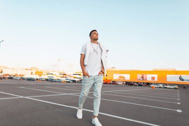 Şık yaz beyaz ve kot giyim bir saç modeli ile deri spor ayakkabı genç yakışıklı adam modeli bir şehir otoparkta yürüyor. Şık Amerikalı adam bir yürüyüş sahiptir. Moda yazlık erkek giyim.