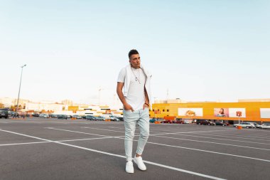 Bir saç modeli ile beyaz şık spor ayakkabı şık bir t-shirt vintage mavi kot trendy genç adam bir yaz gününde şehir etrafında seyahat. Yakışıklı adam model şehir otoparkında yürüyor.