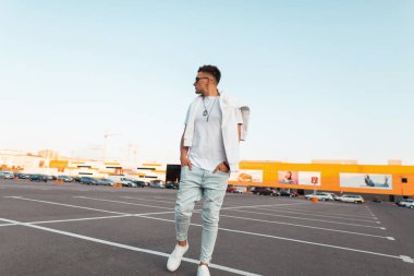 Şık bir kazak ile trendy güneş gözlüğü bir vintage beyaz t-shirt şık kot şık genç hipster adam mağaza yakınındaki otoparkta yürüyor. Çekici modern adam sokakta seyahat.