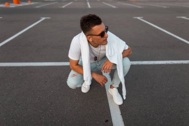 Bir yolda dinleniyor bir sweatshirt ile şık bir t-shirt beyaz spor ayakkabı lı mavi kot pantolon güneş gözlüğü Amerikan genç hipster adam. Yakışıklı şehirli adam otoparkta poz verecek. Şık yaz erkek giyim.