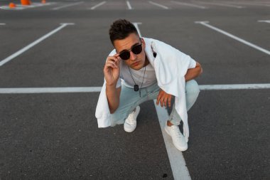 Bir asfalt üzerinde oturan bir sweatshirt ile bir moda t-shirt beyaz spor ayakkabı lı mavi kot pantolon modern genç hipster adam ve kamera bakmak. Serin şehir adam otoparkta rahatlatır. Şık erkek giyim.